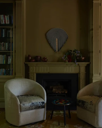 Interior of a living room with two armchairs, a small table, fireplace and mantel, a decorative wall art, bookshelves, and dark green walls.
