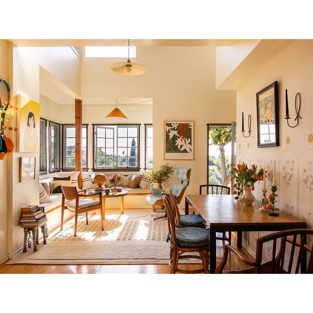Sunlit dining room and living space with a wooden dining table, chairs, a window seat, artwork, and a neutral color palette.