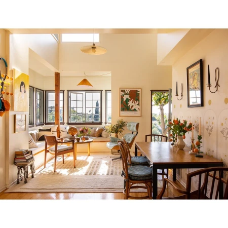 Sunlit dining room and living space with a wooden dining table, chairs, a window seat, artwork, and a neutral color palette.