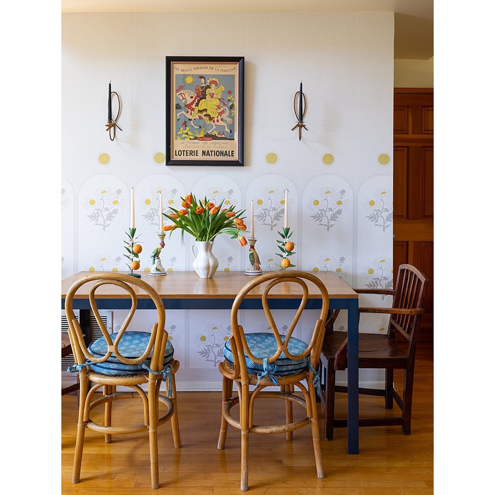 Dining room interior with a wooden table, rattan chairs with blue cushions, white walls with artwork, and a vase of orange tulips.