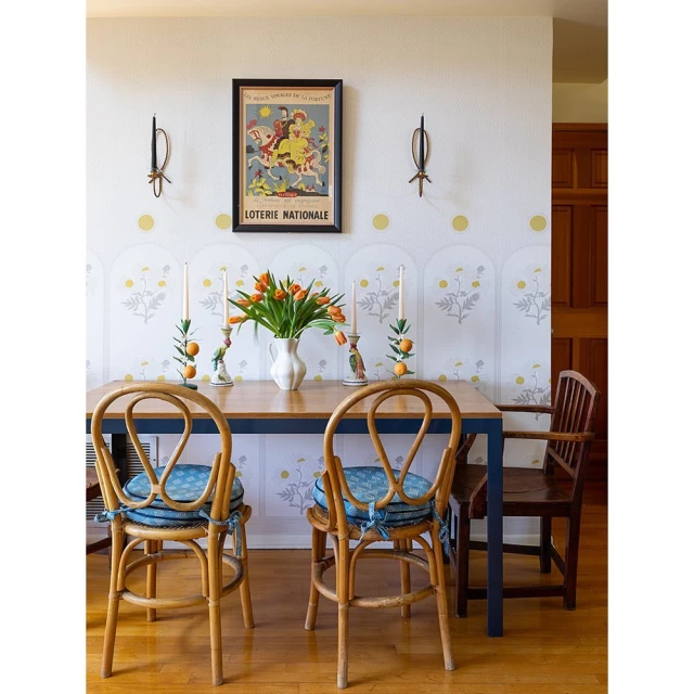 Dining room interior with a wooden table, rattan chairs with blue cushions, white walls with artwork, and a vase of orange tulips.