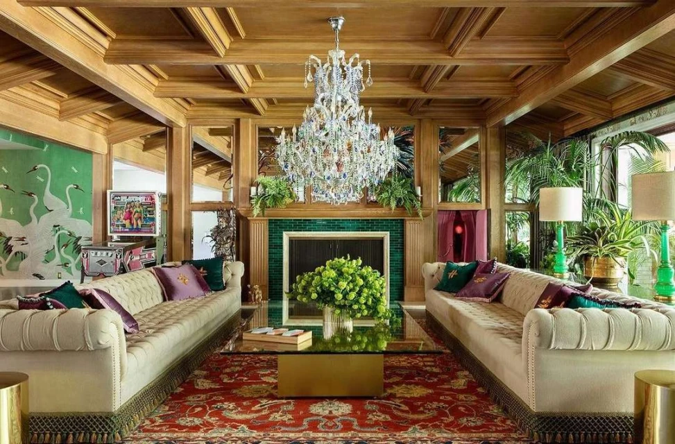 Opulent living room features a crystal chandelier, sofas with throw pillows, and a patterned rug. Houseplants add a touch of nature.