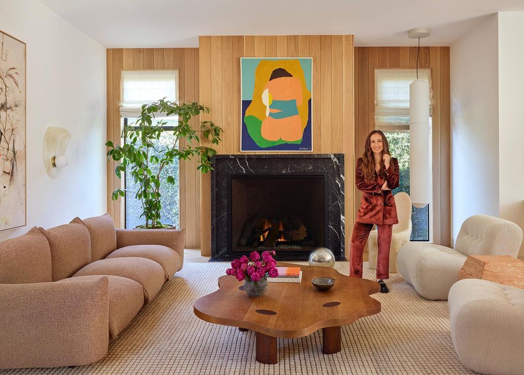 Living room interior featuring an adult woman in a velvet suit, a unique coffee table, and a fireplace with an abstract art piece above.