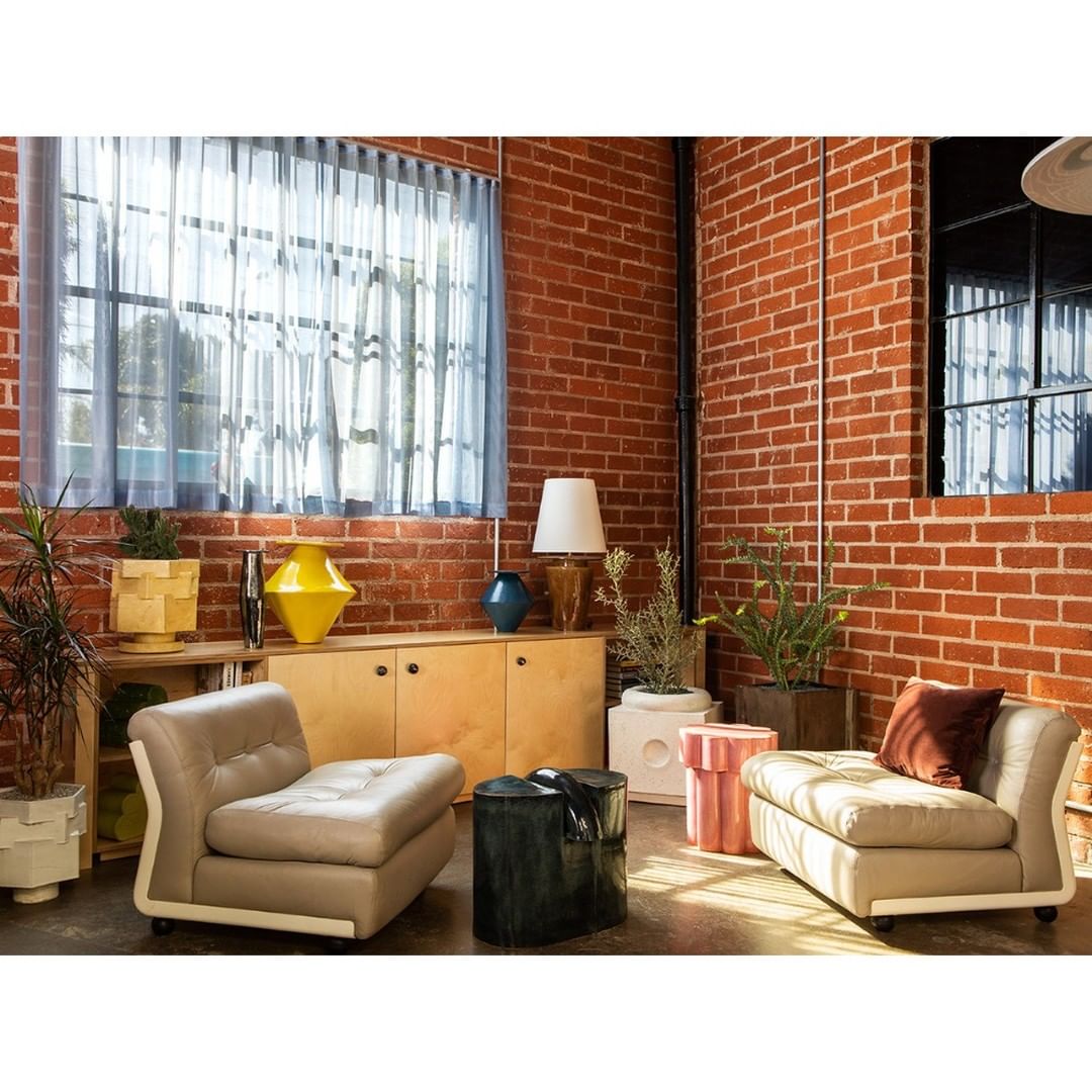 Living room setup with modern beige armchairs, potted plants, and a minimalist wooden cabinet against a textured brick wall backdrop.