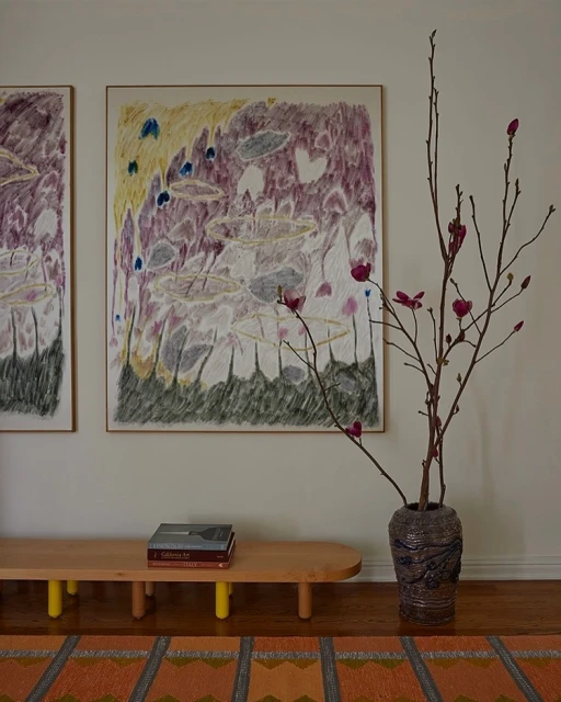 Interior with framed abstract art, a blonde wood bench with books, a vase of floral branches, and a geometric patterned rug on a hardwood floor.