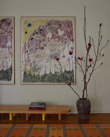 Interior with framed abstract art, a blonde wood bench with books, a vase of floral branches, and a geometric patterned rug on a hardwood floor.