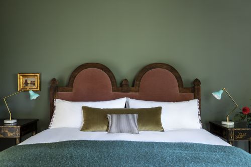 Interior design photograph of a bed with a velvet headboard, white linens, lamps on bedside tables, and an olive green wall.