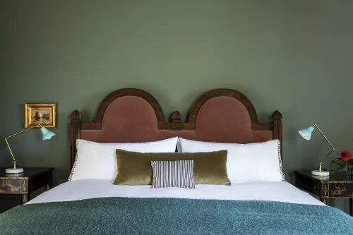 Interior design photograph of a bed with a velvet headboard, white linens, lamps on bedside tables, and an olive green wall.