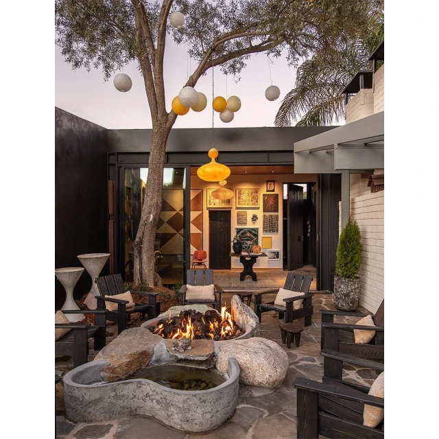 Outdoor living space with a stone fire pit, water feature, wooden chairs, a leafy tree, and hanging pendant lights at dusk.