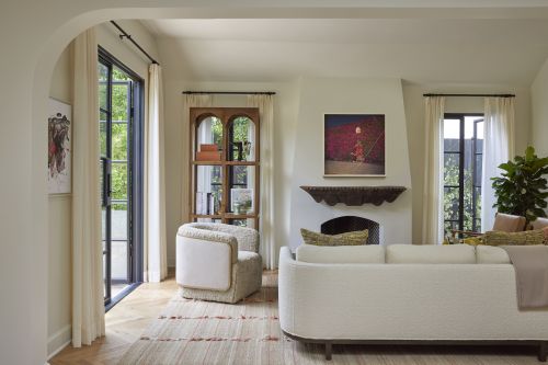 Living room interior with white sofa, armchair, fireplace, and bookshelf, bathed in natural light from windows with sheer white curtains.