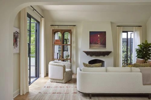 Living room interior with white sofa, armchair, fireplace, and bookshelf, bathed in natural light from windows with sheer white curtains.