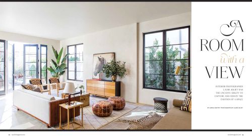 Well-lit living room with a white sofa, wood furniture, decorative pillows, and large windows, offering a view of the outside greenery.