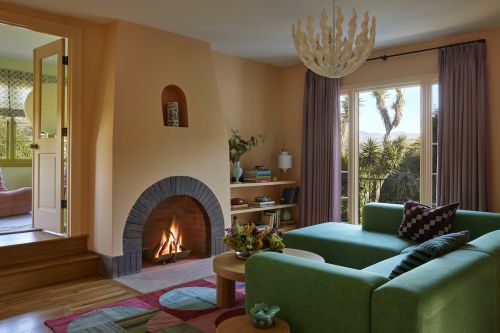 Living room interior features a green sectional sofa, fireplace with brick arch, colorful rug, and natural light from a window view.