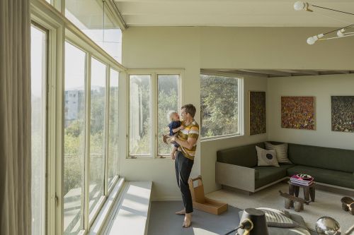 Father holding his baby son in a modern living room near large windows, showcasing a tender family moment with contemporary interior design.