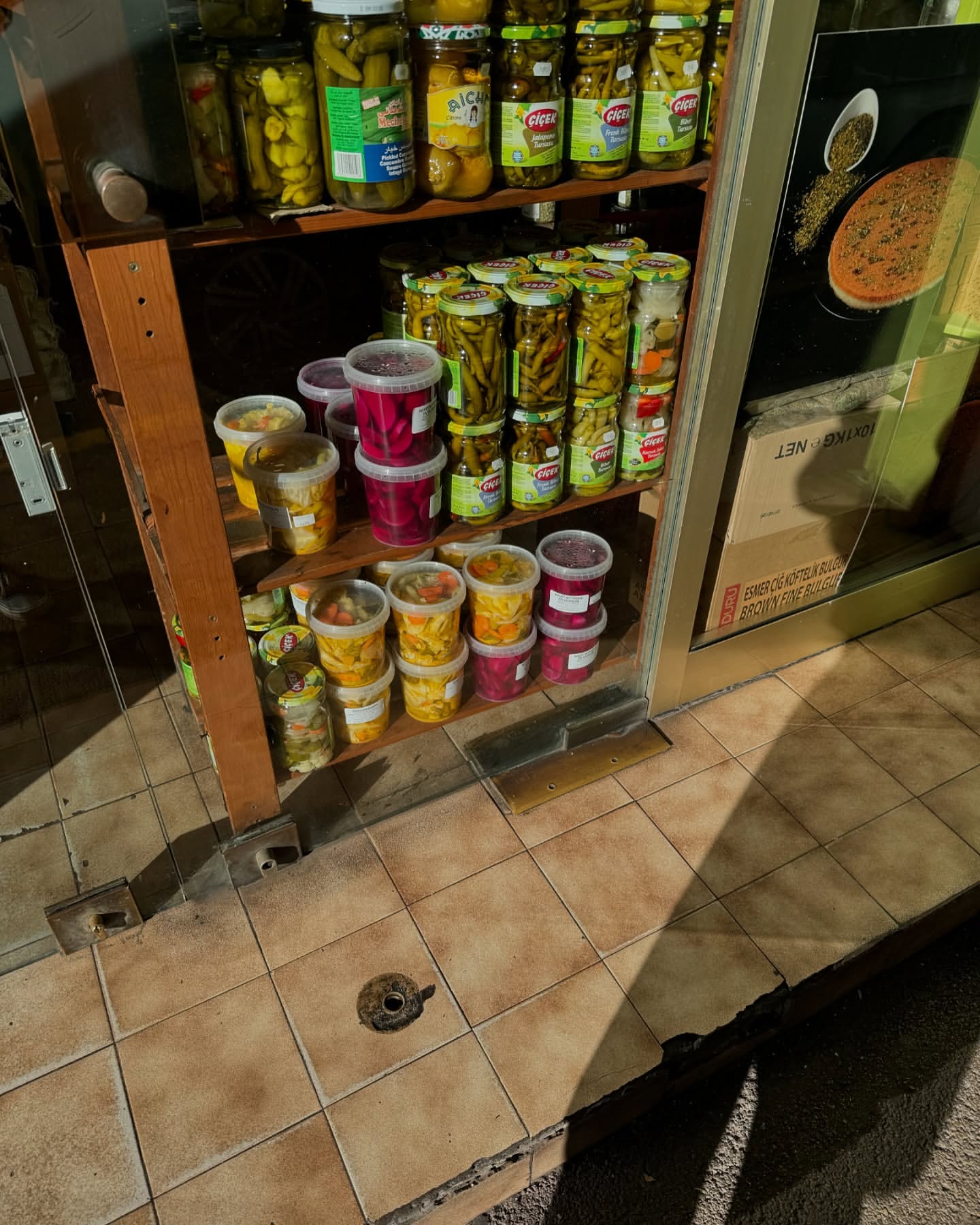 Display of pickled vegetables in jars and plastic containers on wooden shelves, next to a storefront entrance with tiled flooring.