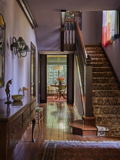 Interior view of a home hallway with a wooden staircase, runner rug, console table with decor, and a glimpse into a dining room.
