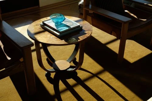 Still life in living room: Round wood table with blue glass bowl and books, next to tan armchair and shadows on gold carpet.