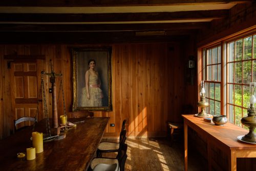 Interior of a wood-paneled room featuring a painting of a woman, antique furniture, a long table, and natural light streaming through windows.