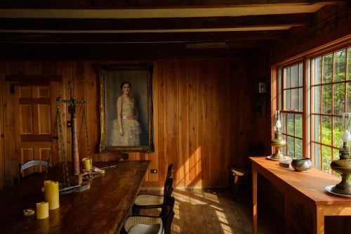 Interior of a wood-paneled room featuring a painting of a woman, antique furniture, a long table, and natural light streaming through windows.