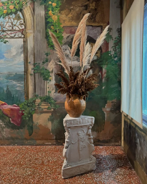 Floral arrangement featuring pampas grass and dried flowers in a vase on a pedestal against a mural backdrop.
