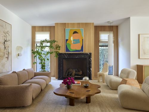 Living room interior with a beige sofa, wooden coffee table with pink flowers, and artwork above the fireplace, creating a cozy, inviting space.