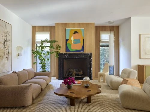 Living room interior with a beige sofa, wooden coffee table with pink flowers, and artwork above the fireplace, creating a cozy, inviting space.