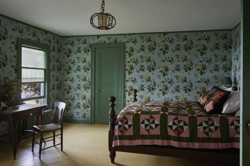 Bedroom interior features a bed with a quilt, floral wallpaper, a wooden desk, and a chair, capturing a cozy and vintage aesthetic.