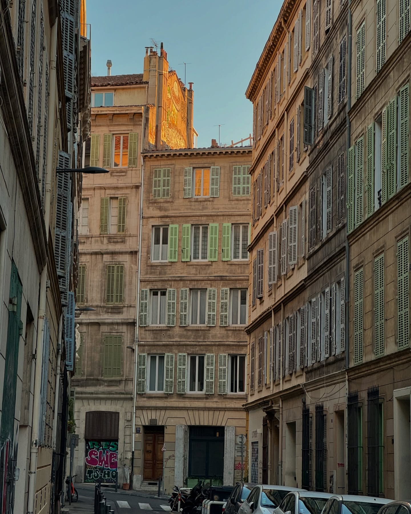Urban street scene in Europe featuring old apartment buildings with faded facades and weathered shutters in shades of green and brown.