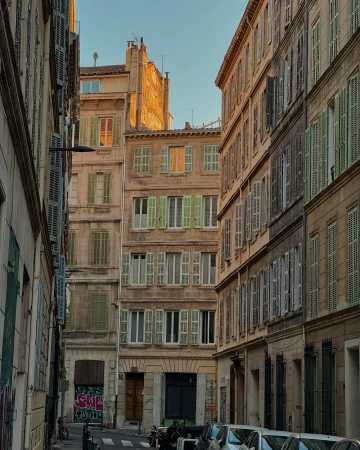 Urban street scene in Europe featuring old apartment buildings with faded facades and weathered shutters in shades of green and brown.