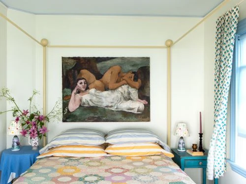 Bedroom interior featuring a painting of reclining figures above a bed with floral bedding, complemented by patterned curtains and soft lighting.
