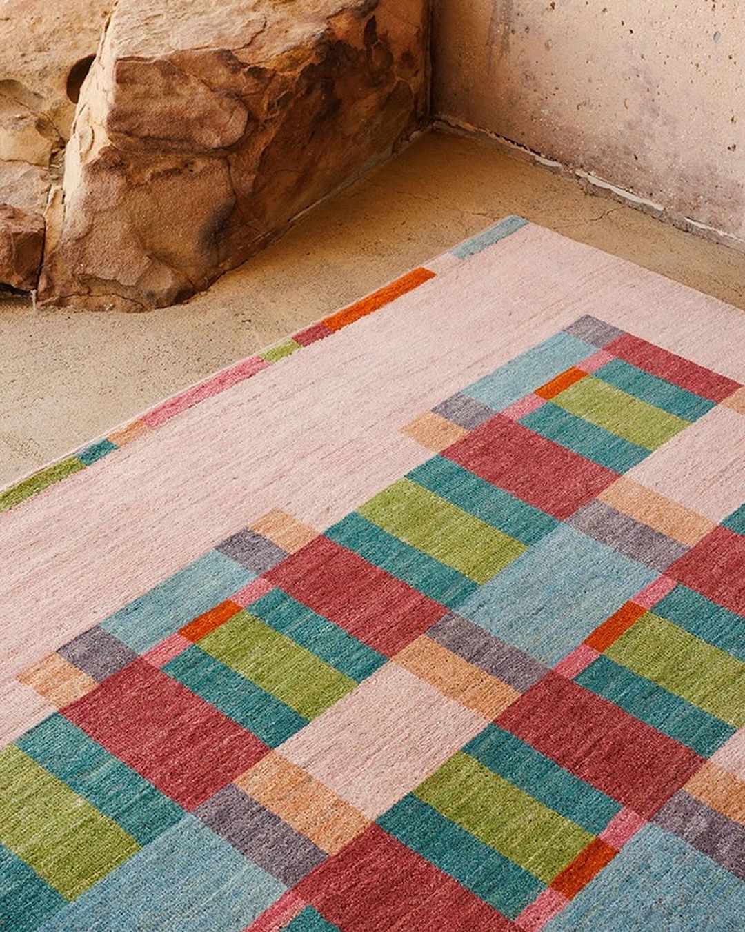 High-angle shot of a colorful geometric rug with blue, green, red, and orange squares, resting on a concrete floor near a sandstone block.
