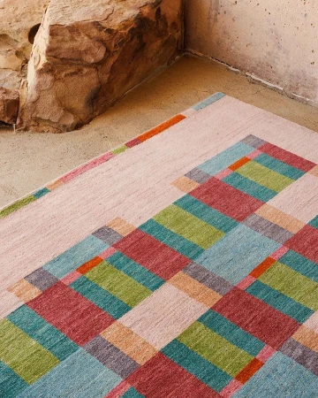 High-angle shot of a colorful geometric rug with blue, green, red, and orange squares, resting on a concrete floor near a sandstone block.