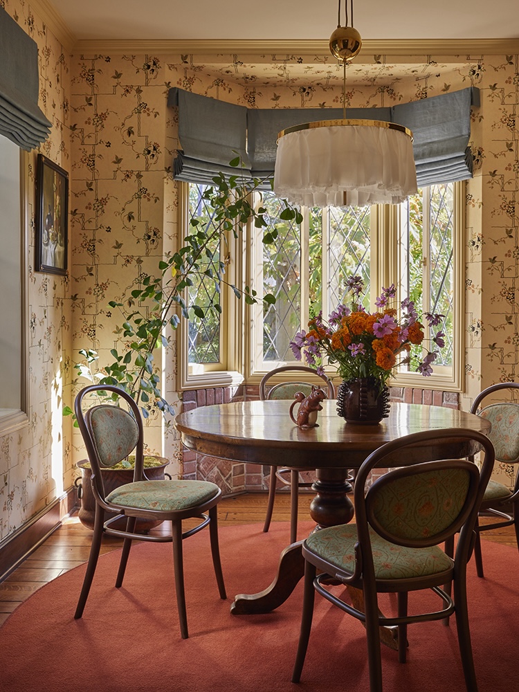 Dining room interior featuring a round table, four chairs, a floral arrangement, and a large window with natural light.