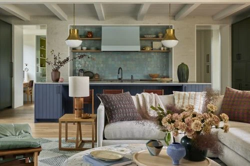 Living room interior with a view into the kitchen, featuring neutral toned sofa, blue cabinets, and modern pendant lighting.