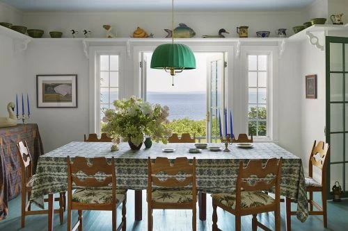 Interior of a dining room with a table set for a meal, wooden chairs, a green chandelier, and a view of the ocean through open windows.