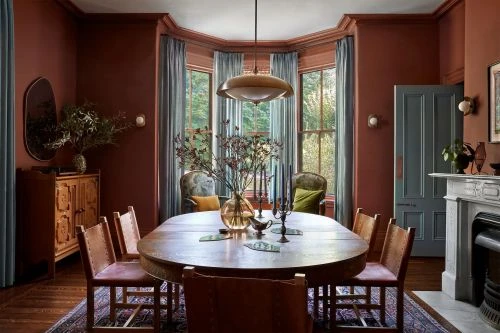 Warm-toned dining room interior featuring a wooden table set with decor, surrounded by leather chairs, with natural light from windows.