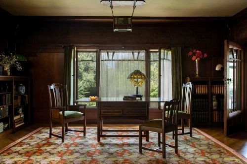 Interior view of a dining room with wooden chairs and table, lit by a stained glass lamp and natural light from a window.