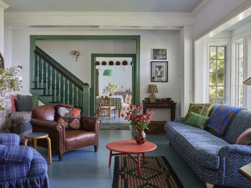 Living room interior with a blue patterned sofa, a leather armchair, a coffee table, and stairs in the background, bathed in natural light.