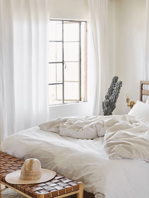 Bright airy bedroom interior featuring a large bed with white linens, a woven bench, a straw hat, and a large window with white curtains.