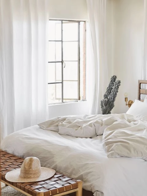 Bright airy bedroom interior featuring a large bed with white linens, a woven bench, a straw hat, and a large window with white curtains.