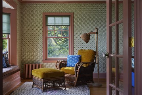 Interior with patterned wallpaper, sunlight through the window, wicker chair and ottoman with soft yellow cushions, and a floor lamp.