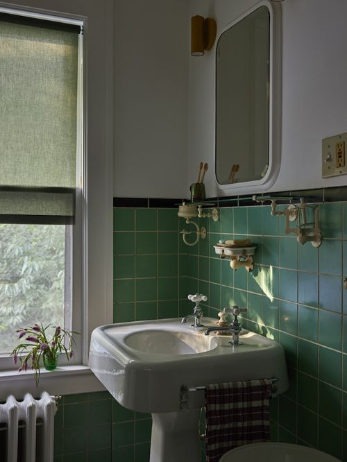 Bathroom interior featuring a pedestal sink with green tiled walls, a window with a green shade, and a white framed mirror.
