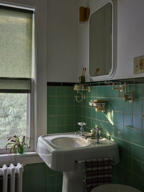 Bathroom interior featuring a pedestal sink with green tiled walls, a window with a green shade, and a white framed mirror.