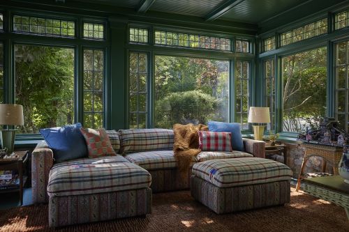 Sunroom interior featuring a plaid patterned sectional couch, plush pillows, lamps, large windows, and a view of a green, leafy outdoor space.