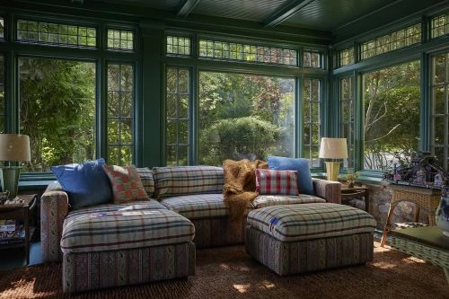Sunroom interior featuring a plaid patterned sectional couch, plush pillows, lamps, large windows, and a view of a green, leafy outdoor space.