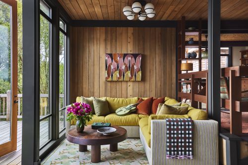 Living room interior with a yellow sectional sofa, wood-paneled walls, large windows, and round coffee table with floral arrangement.