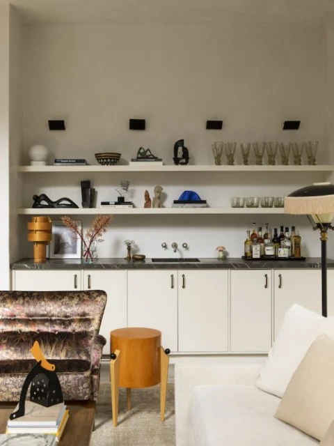 Interior view features a decorative shelf with various art objects, glasses, and bottles, with an upholstered chair and end table.