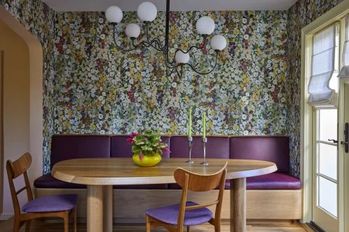 Dining room features a light wood table and chairs, a purple upholstered bench against a floral wallpapered wall, and a modern chandelier.