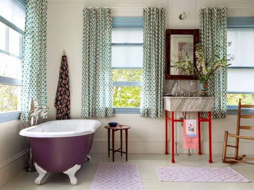 Bathroom interior featuring a purple clawfoot bathtub, vanity with marble countertop, floral drapes, and sunlight streaming through the windows.