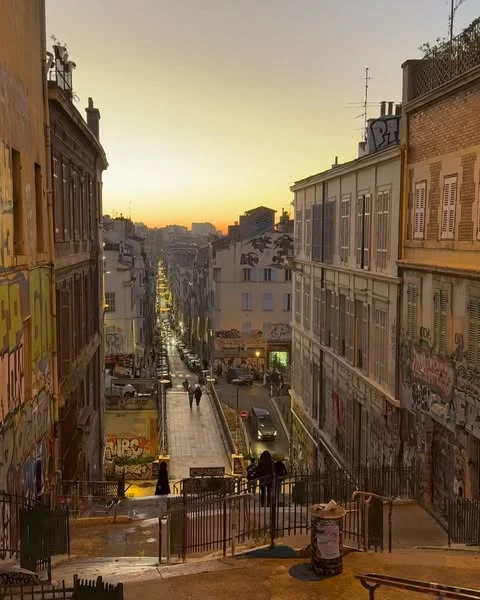 City street scene at dusk featuring apartment buildings with graffiti art, cars on a street, and people walking on the sidewalks.